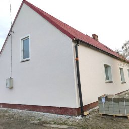 Stanisław Cupryś Stasiu usługi budowlane elewacje - Otynkowany dom jednorodzinny z czerwonym dachem, widok z boku. Na pierwszym planie paleta z bloczkami. Świeżo pomalowana elewacja w jasnym odcieniu.