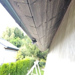 Elewacje Ocieplenia DomówTynki Natryskowe Koszalin, Kołobrzeg SUB 177 Budowlane - Drewniana podbitka dachowa w ciemnym odcieniu, zamontowana przy elewacji budynku. Widoczne detale montażu i fragment rusztowania.