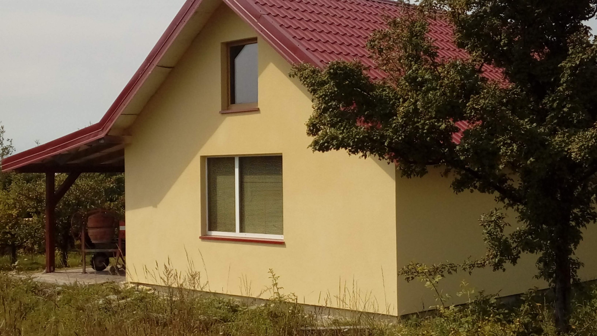 Żółty dom z czerwoną dachówką i białymi ramami okiennymi, częściowo zasłonięty drzewem. Z lewej strony widoczna wiata i betoniarka. Nierówny teren z trawą.
