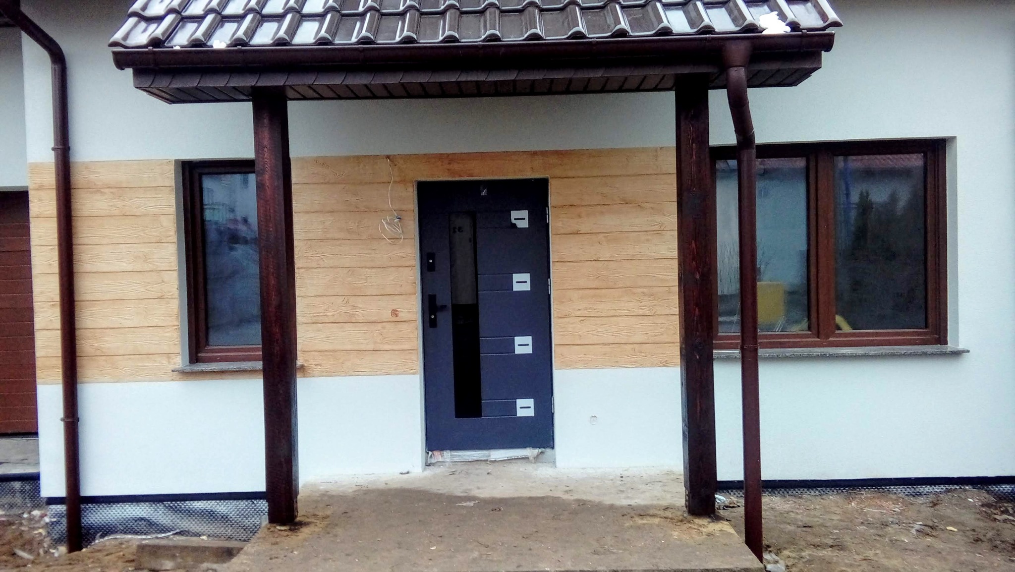 Wykończone wejście do domu z drewnopodobną elewacją, brązowymi oknami i grafitowymi drzwiami z przeszkleniami. Widoczne rynny i słupy podtrzymujące daszek.