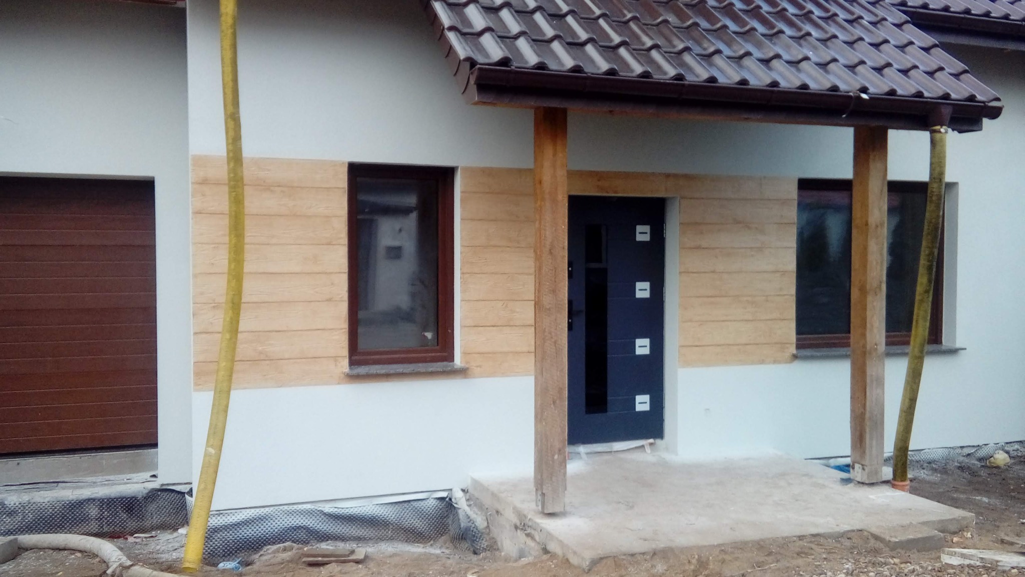 Dom jednorodzinny z elewacją w dwóch odcieniach: jasny beż i imitacja drewna. Widoczna stolarka okienna w brązowych ramach i ciemne drzwi wejściowe.