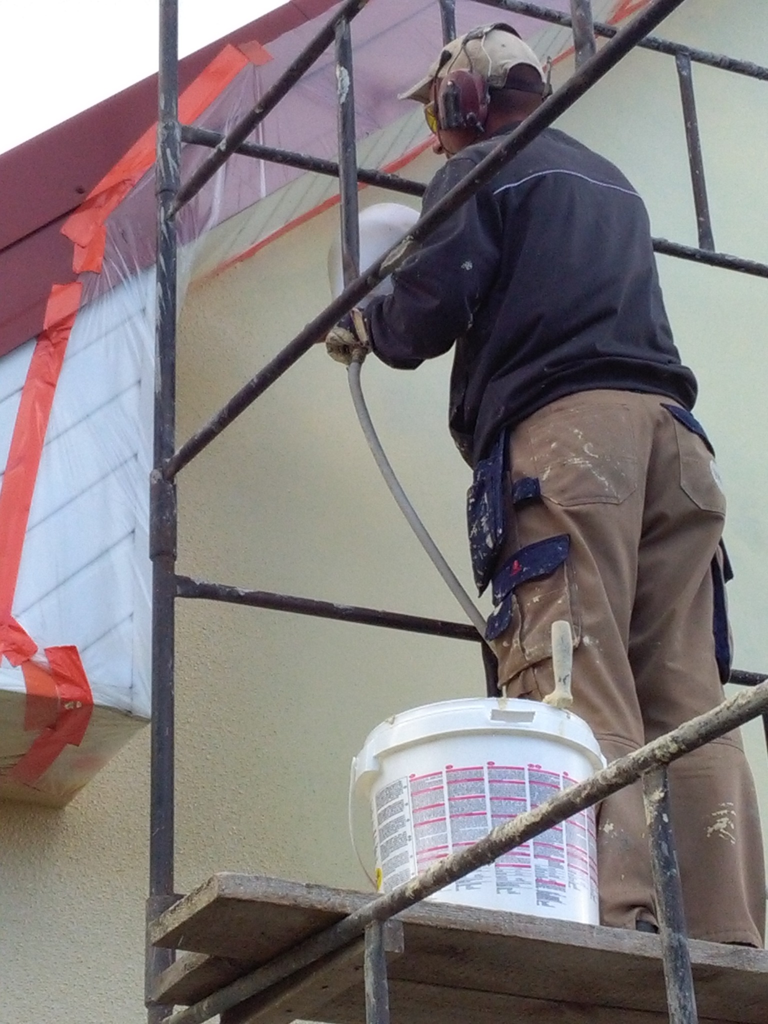 Pracownik na rusztowaniu natryskowo aplikuje preparat na elewację budynku, zabezpieczonego folią i taśmą malarską. Widoczny biały pojemnik z preparatem.