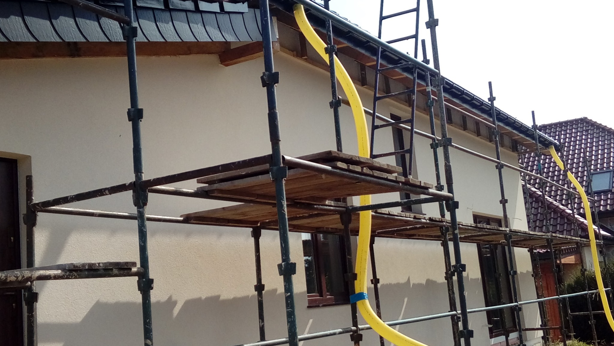 Dom w rusztowaniach z żółtą rurą wentylacyjną. Ściana pokryta jasnym tynkiem, widoczne okna i fragment dachu z ciemnej dachówki. Prace na wysokości.