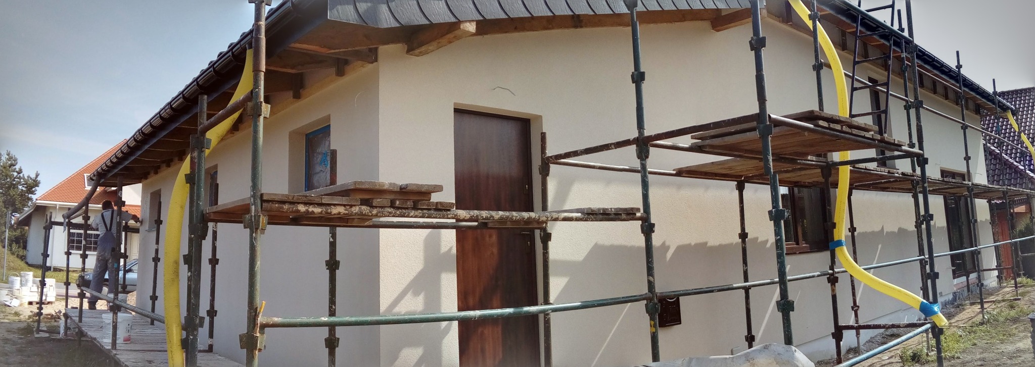 Dom w trakcie prac elewacyjnych, widoczne rusztowania i pracownik. Ściany otynkowane na jasny kolor. Widoczne elementy drewniane i okna zabezpieczone folią.