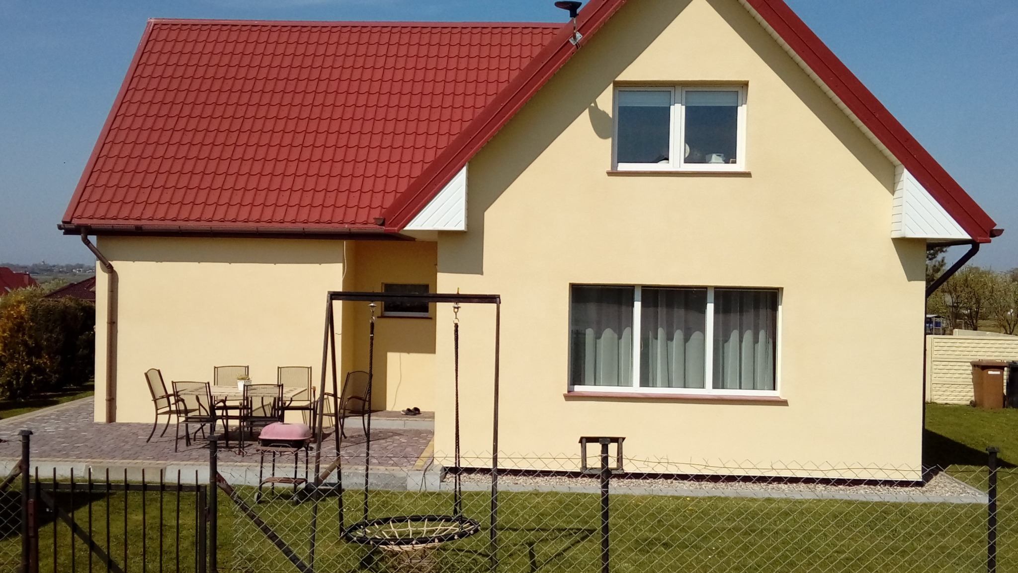 Żółty dom jednorodzinny z czerwoną dachówką, widok zza ogrodzenia. Na tarasie zestaw mebli ogrodowych. Słoneczny dzień, błękitne niebo.