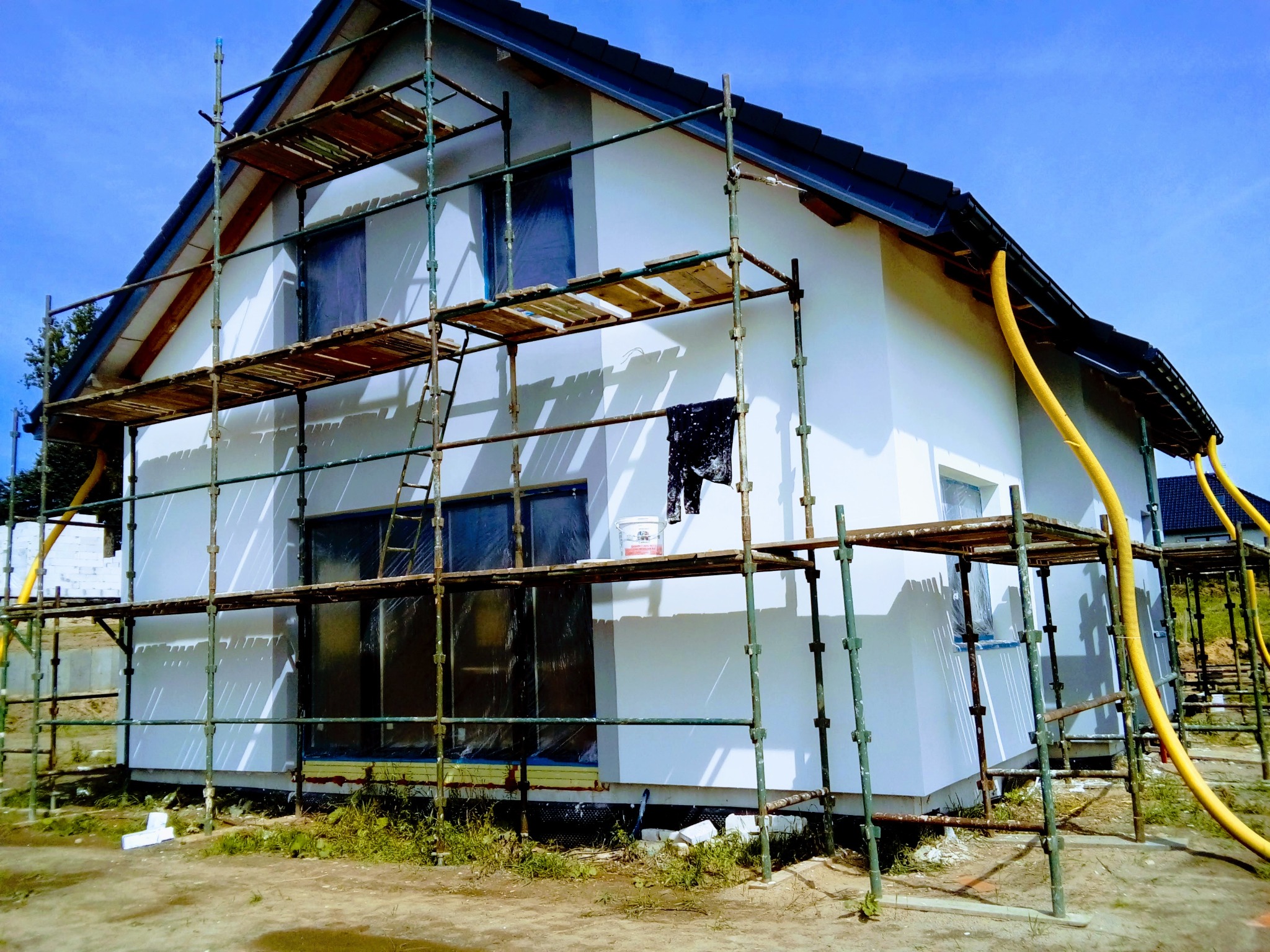 Świeżo pomalowany dom jednorodzinny w rusztowaniach, z żółtymi rurami wentylacyjnymi wzdłuż dachu, okna zabezpieczone folią, słoneczny dzień.