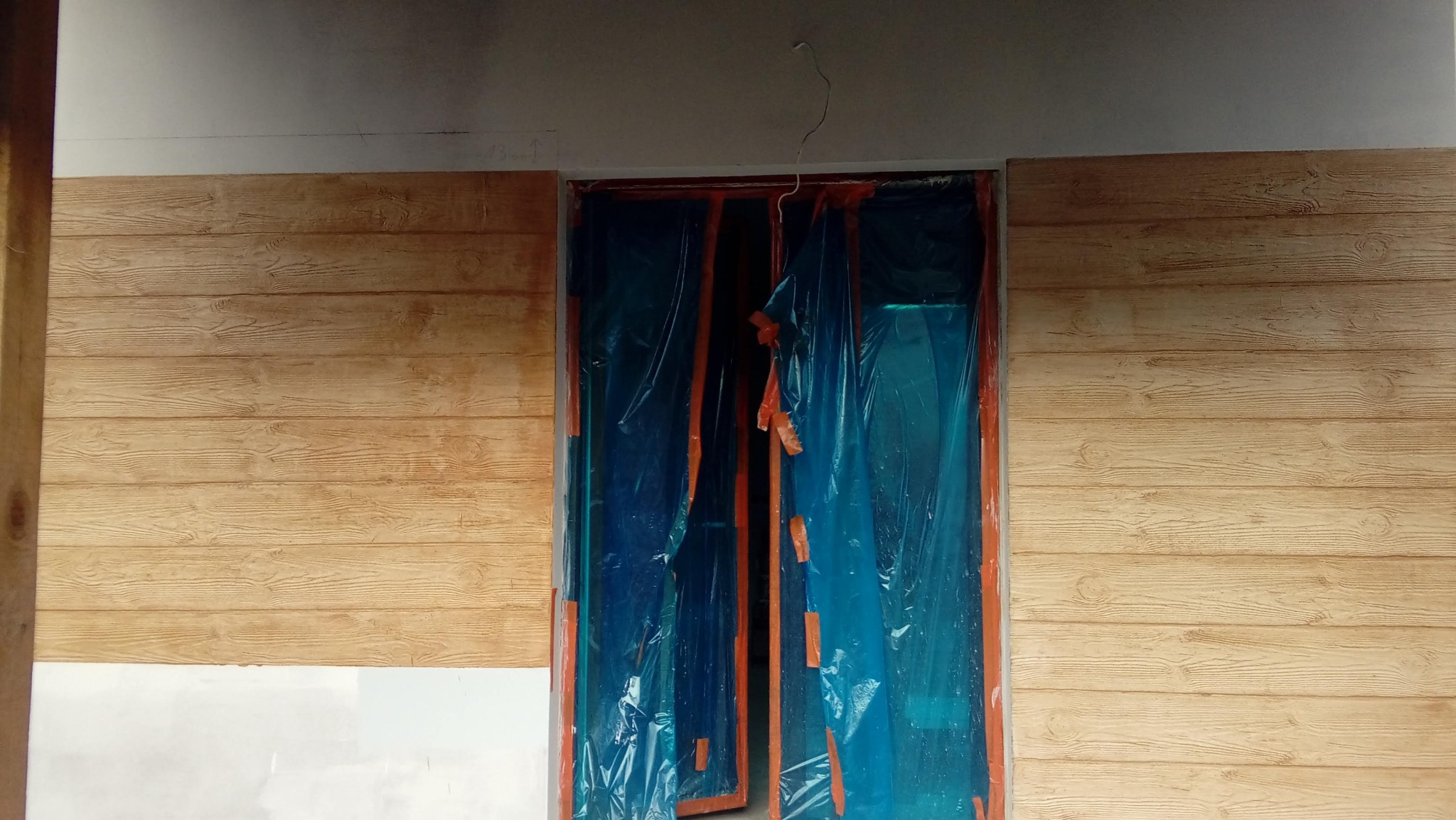 Ściana z efektem drewna, zabezpieczone folią drzwi. Imitacja desek na ścianie, detale słojów widoczne. Prace malarskie w toku, ochrona przed zabrudzeniem.