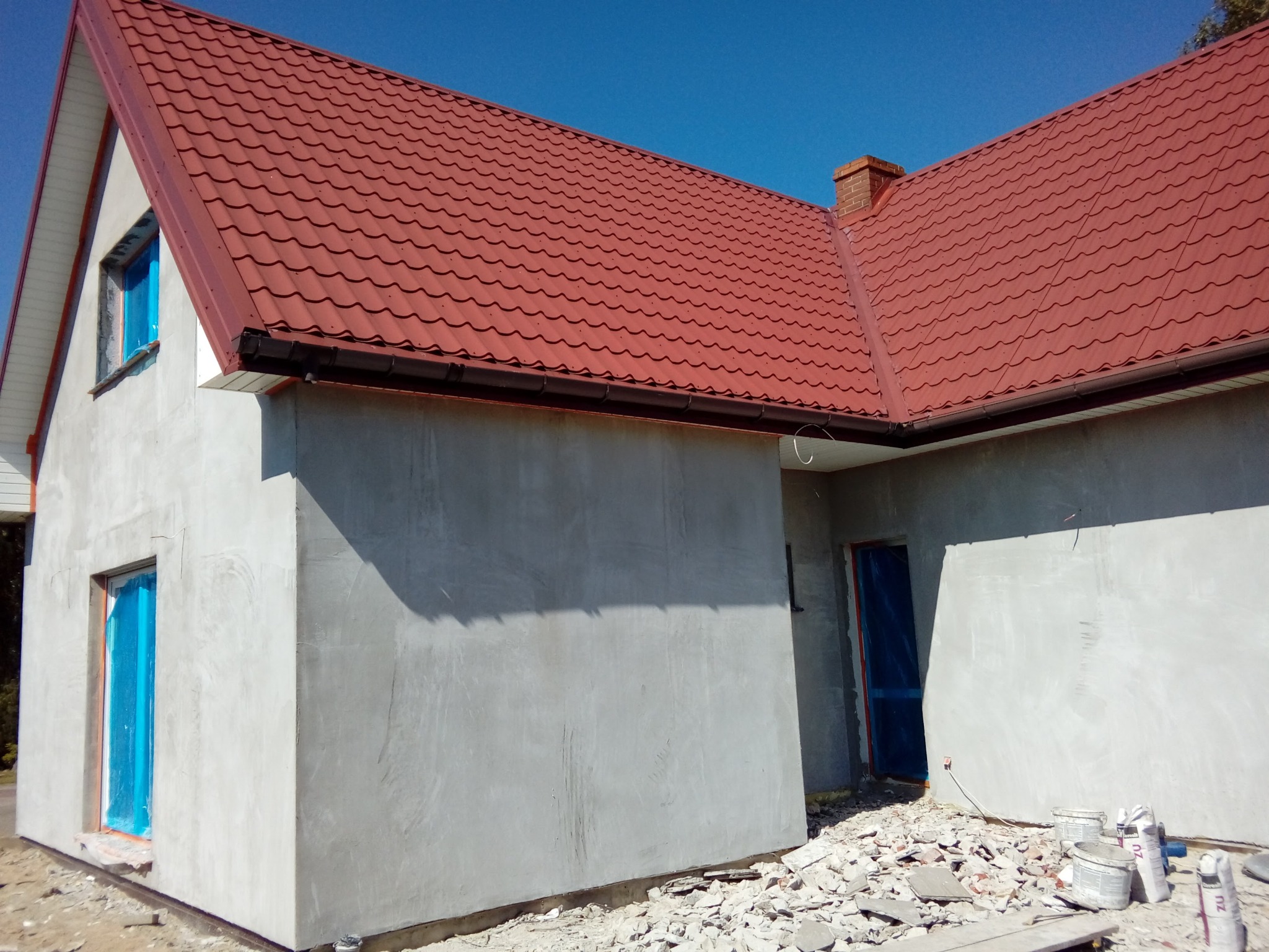 Dom w trakcie prac elewacyjnych: surowe ściany, zabezpieczone okna, czerwony dach. Widoczne gruz i puste wiadra po materiałach budowlanych.