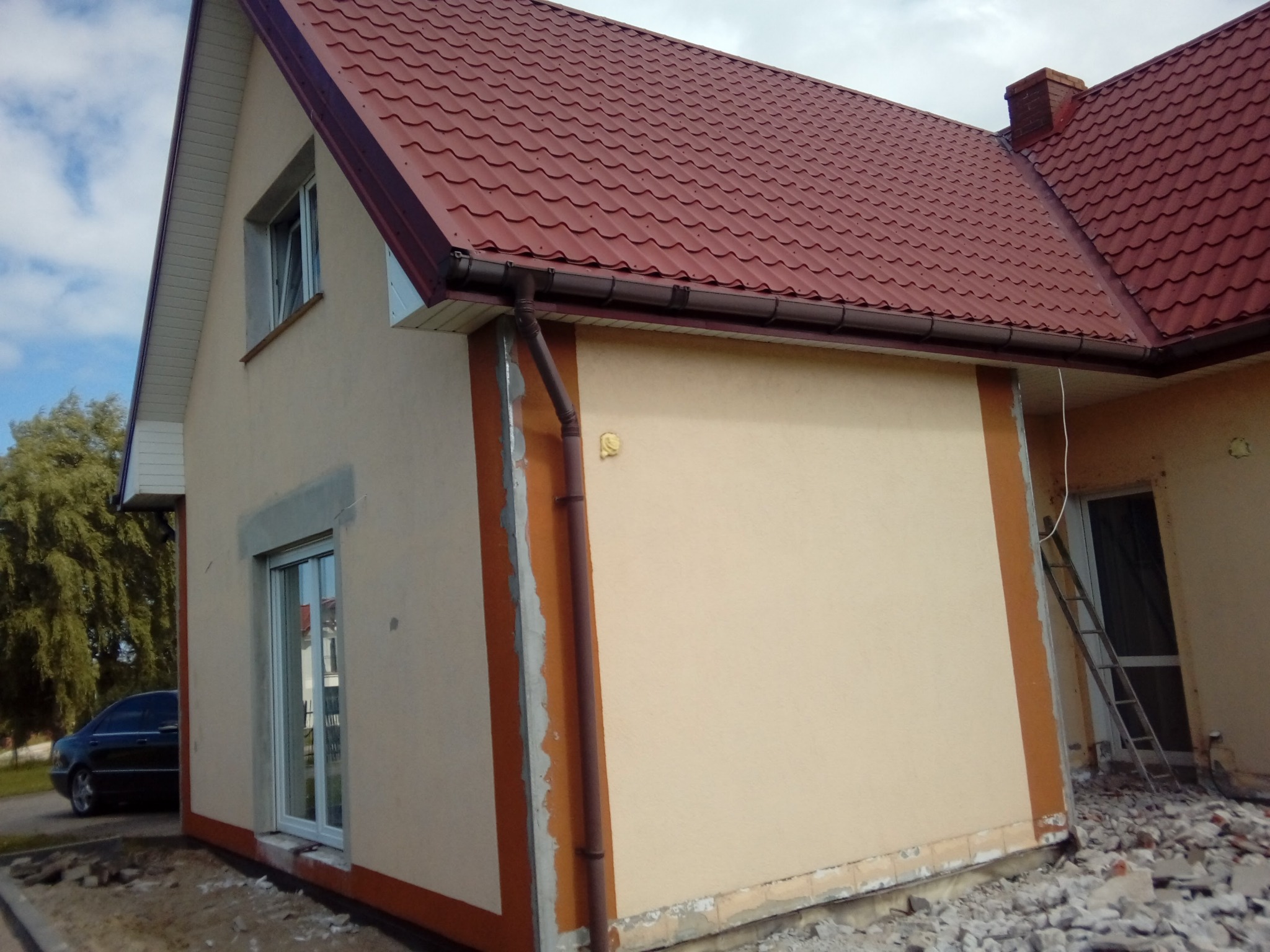 Dom jednorodzinny z nową, beżową elewacją i czerwoną dachówką. Widoczne fragmenty elewacji w trakcie wykończenia, okna z szarymi obramowaniami.