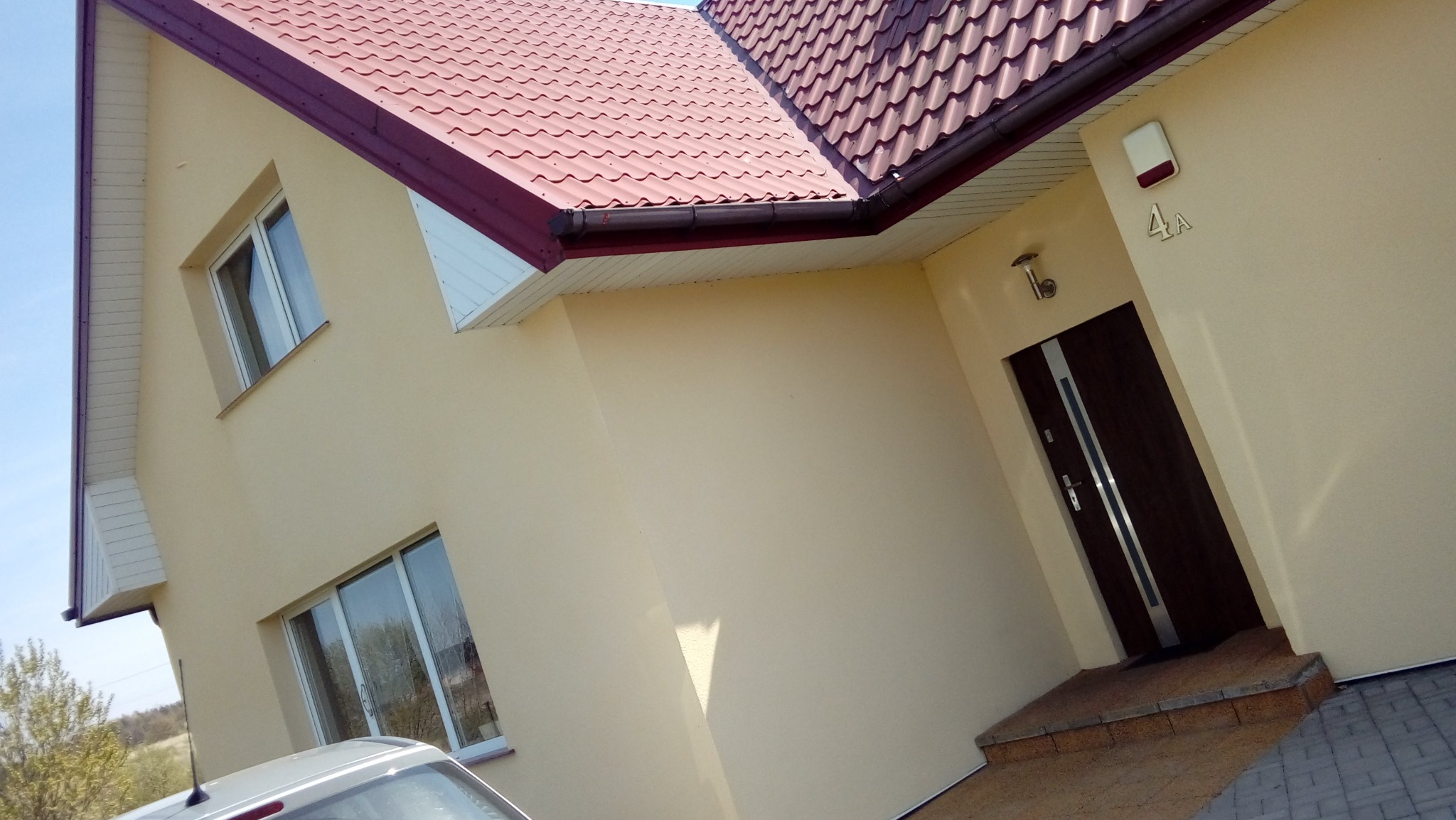 Jasna elewacja domu jednorodzinnego z ciemnobrązowymi oknami i drzwiami wejściowymi, czerwony dach, widok na fragment samochodu.