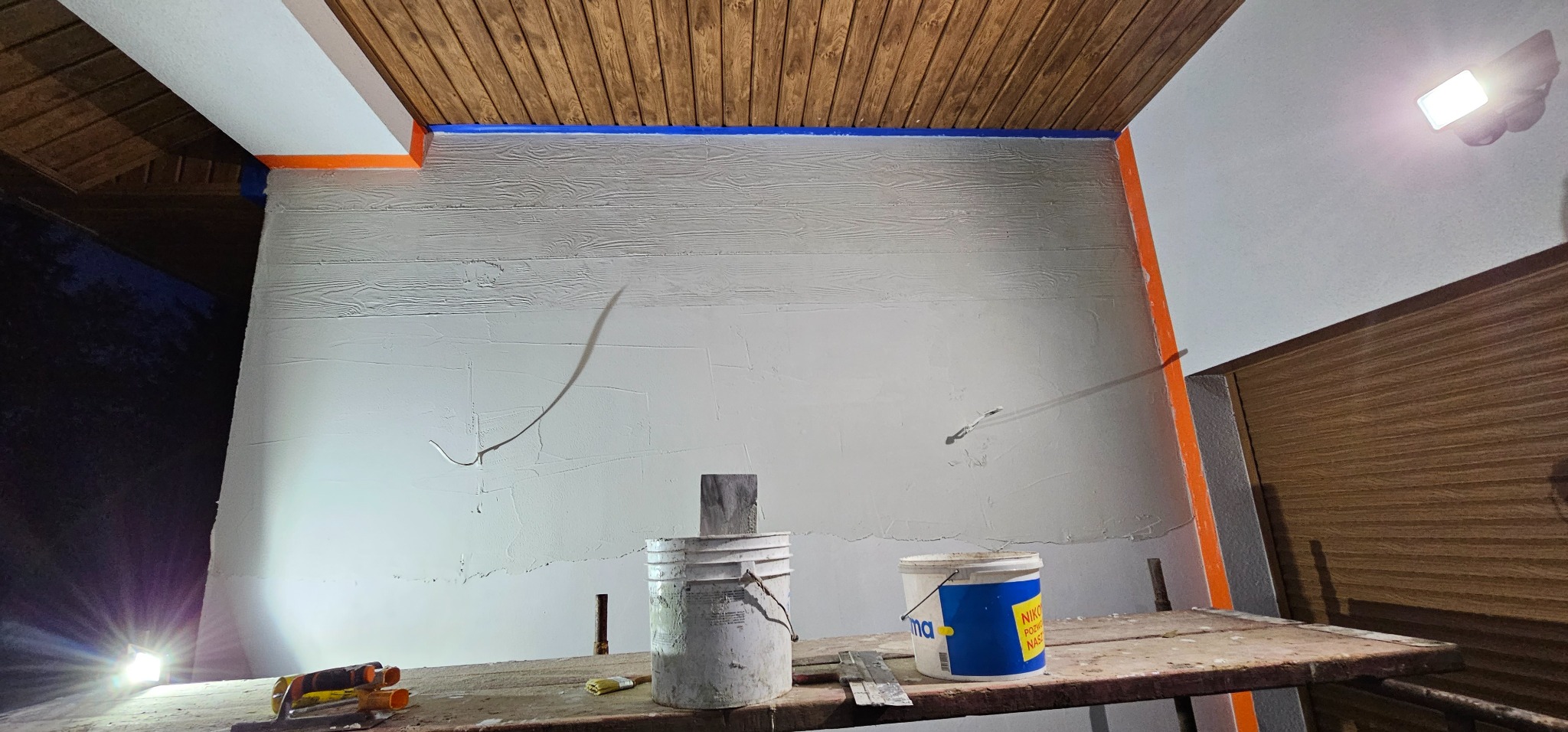 Ściana z nałożoną fakturą imitującą drewno, zabezpieczona taśmą malarską. Na rusztowaniu wiadra z farbą i narzędzia. Oświetlenie robocze podkreśla strukturę.