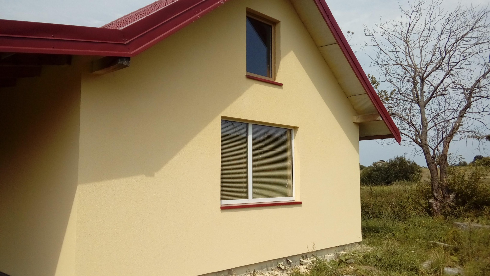 Jasnożółty dom z czerwonym dachem i białymi oknami. Widoczna faktura elewacji, zieleń w tle i fragment nagiego drzewa. Ujęcie z boku, w słoneczny dzień.