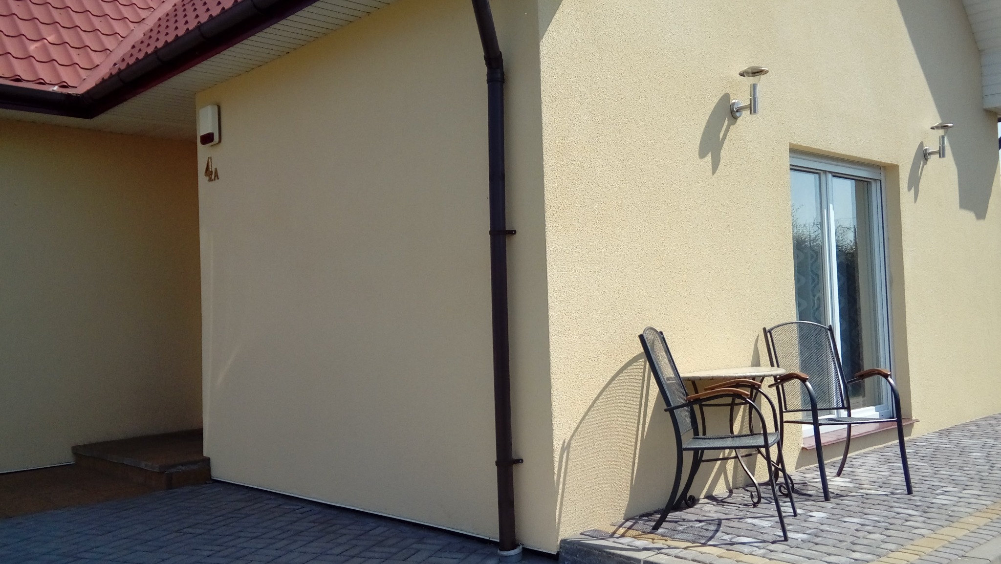 Jasna elewacja domu z numerem '4a', brązową rynną i lampami. Na zewnątrz stoi metalowy stolik z krzesłami. Kostka brukowa na podłożu.