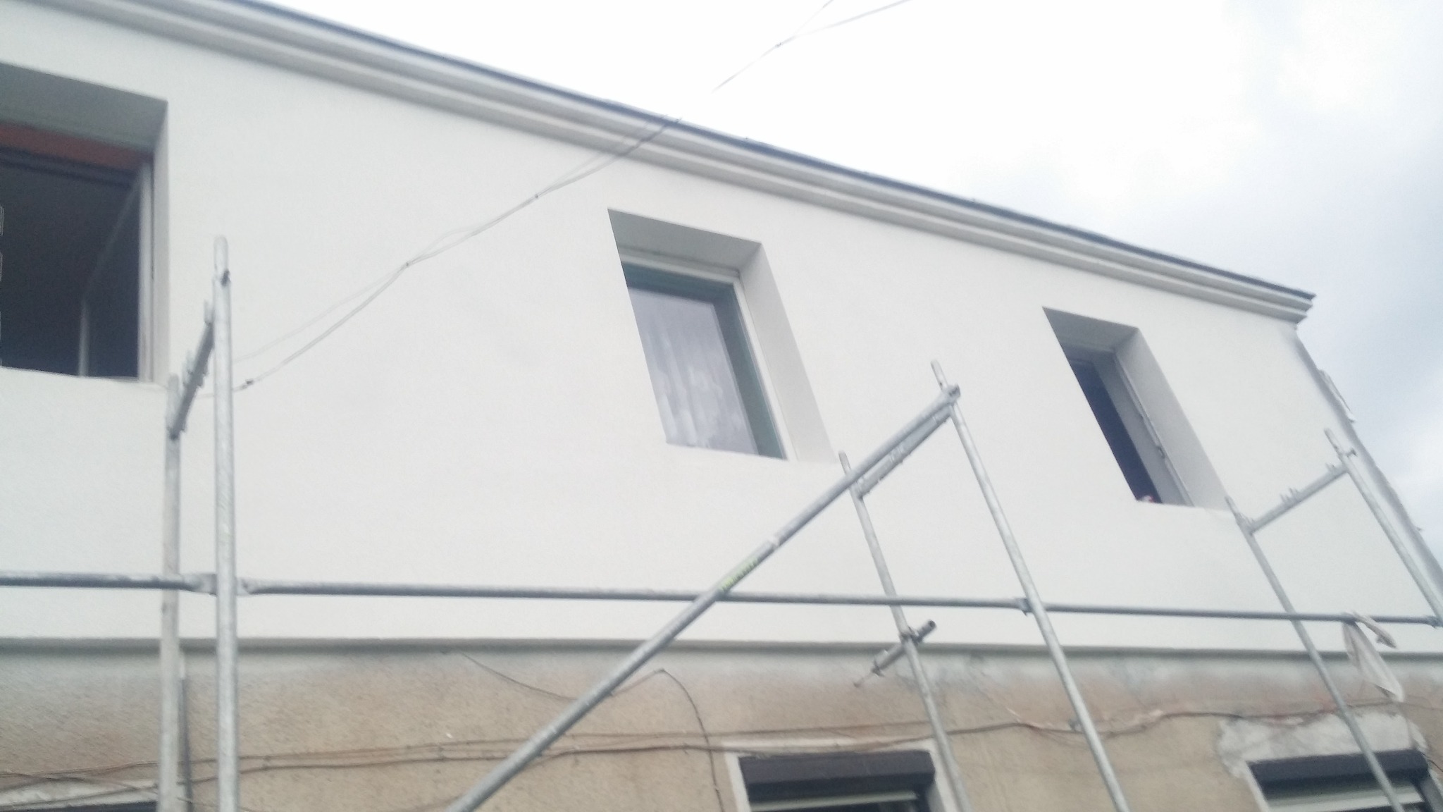 Biała elewacja budynku po renowacji, widoczna z dołu, z oknami i fragmentem rusztowania. Górna część ściany pomalowana, dolna w trakcie prac.