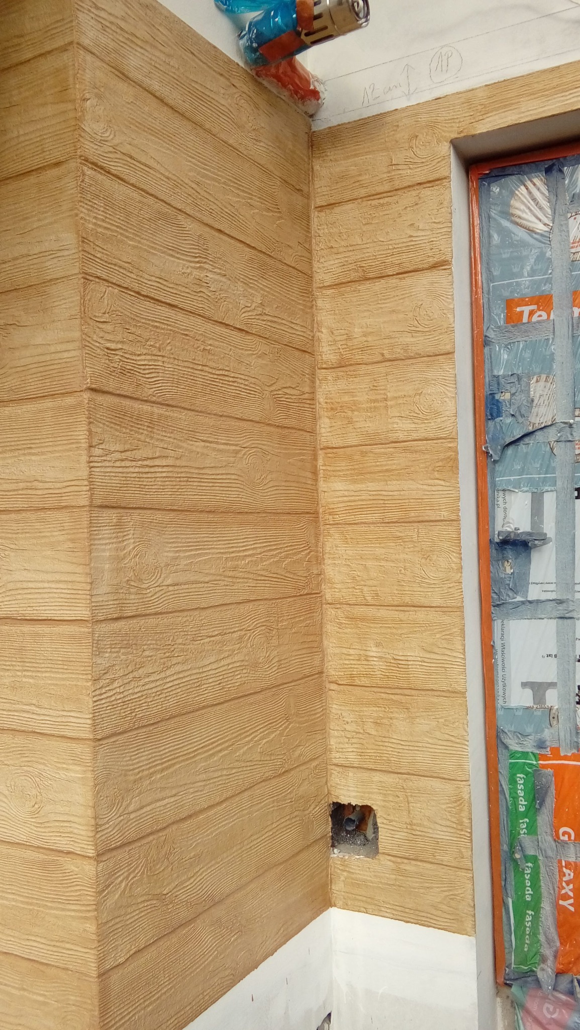 Elewacja budynku z fakturą drewna, zabezpieczone okno folią malarską. Widoczny narożnik i fragment instalacji. Ciepłe, naturalne odcienie brązu i beżu.