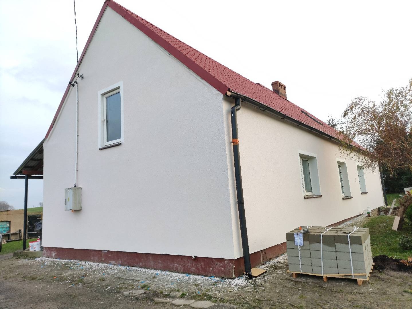 Otynkowany dom jednorodzinny z czerwonym dachem, widok z boku. Na pierwszym planie paleta z bloczkami. Świeżo pomalowana elewacja w jasnym odcieniu.
