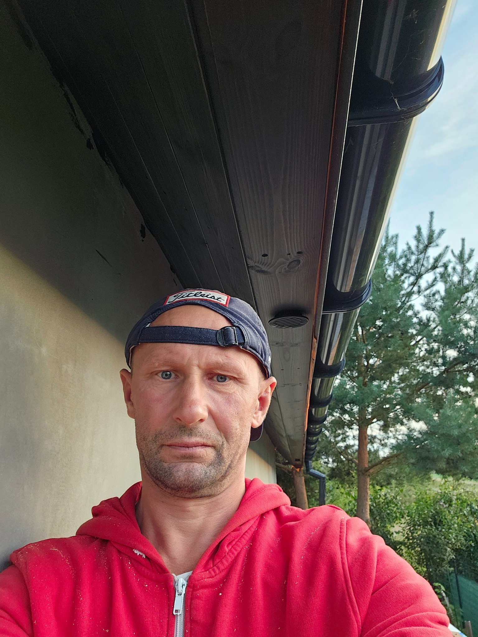 Mężczyzna w czerwonej bluzie i czapce na tle elewacji z zamontowaną, czarną rynną. Widoczne drzewa w tle. Ujęcie z dołu, podkreślające detale rynny.