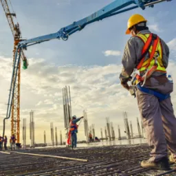 Praca na budowie: robotnicy w kaskach, zbrojenie, kolumny, żuraw do betonu na tle nieba. Widok z dołu, perspektywa budowlana, etap wylewania fundamentów.