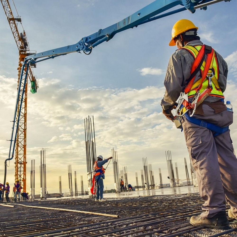 Praca na budowie: robotnicy w kaskach, zbrojenie, kolumny, żuraw do betonu na tle nieba. Widok z dołu, perspektywa budowlana, etap wylewania fundamentów.