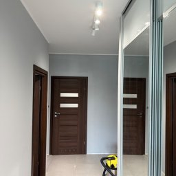 M&D Remont4everyone Mykola Petruniv - Szaro-białe wnętrze z ciemnymi drzwiami i przesuwną szafą z lustrami. Na podłodze widoczny odkurzacz i klapki. Świeżo po remoncie.