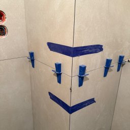 M&D Remont4everyone Mykola Petruniv - Układanie jasnych płytek w narożniku z użyciem niebieskiej taśmy i klipsów poziomujących we Wrocławiu. Widoczne gniazdka elektryczne w ścianie.