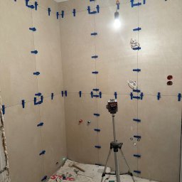 M&D Remont4everyone Mykola Petruniv - Układanie płytek w łazience z użyciem poziomicy laserowej i systemu poziomowania płytek. Widoczne narzędzia i materiały budowlane. Jasne płytki na ścianach.