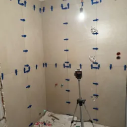 Układanie płytek w łazience z użyciem poziomicy laserowej i systemu poziomowania płytek. Widoczne narzędzia i materiały budowlane. Jasne płytki na ścianach.