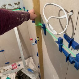 M&D Remont4everyone Mykola Petruniv - Układanie płytek na ścianie z użyciem poziomicy laserowej i klipsów. Widoczny fragment ręki w swetrze, wiadro z klejem i narzędzia glazurnicze.