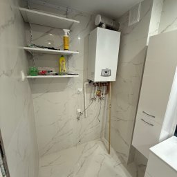 M&D Remont4everyone Mykola Petruniv - Biała łazienka z marmurowymi płytkami, wiszącym piecem gazowym Termet, półkami i szafką pod umywalką. Widoczne rury i instalacja. Nowoczesny design.