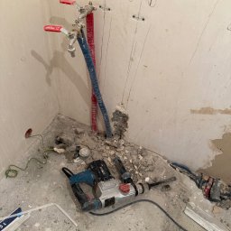 M&D Remont4everyone Mykola Petruniv - Remont łazienki w trakcie: instalacja wodna z zaworami, kable elektryczne, ślady po kuciu w ścianie, gruz i wiertarka udarowa na podłodze. Widok na narożnik pomieszczenia.