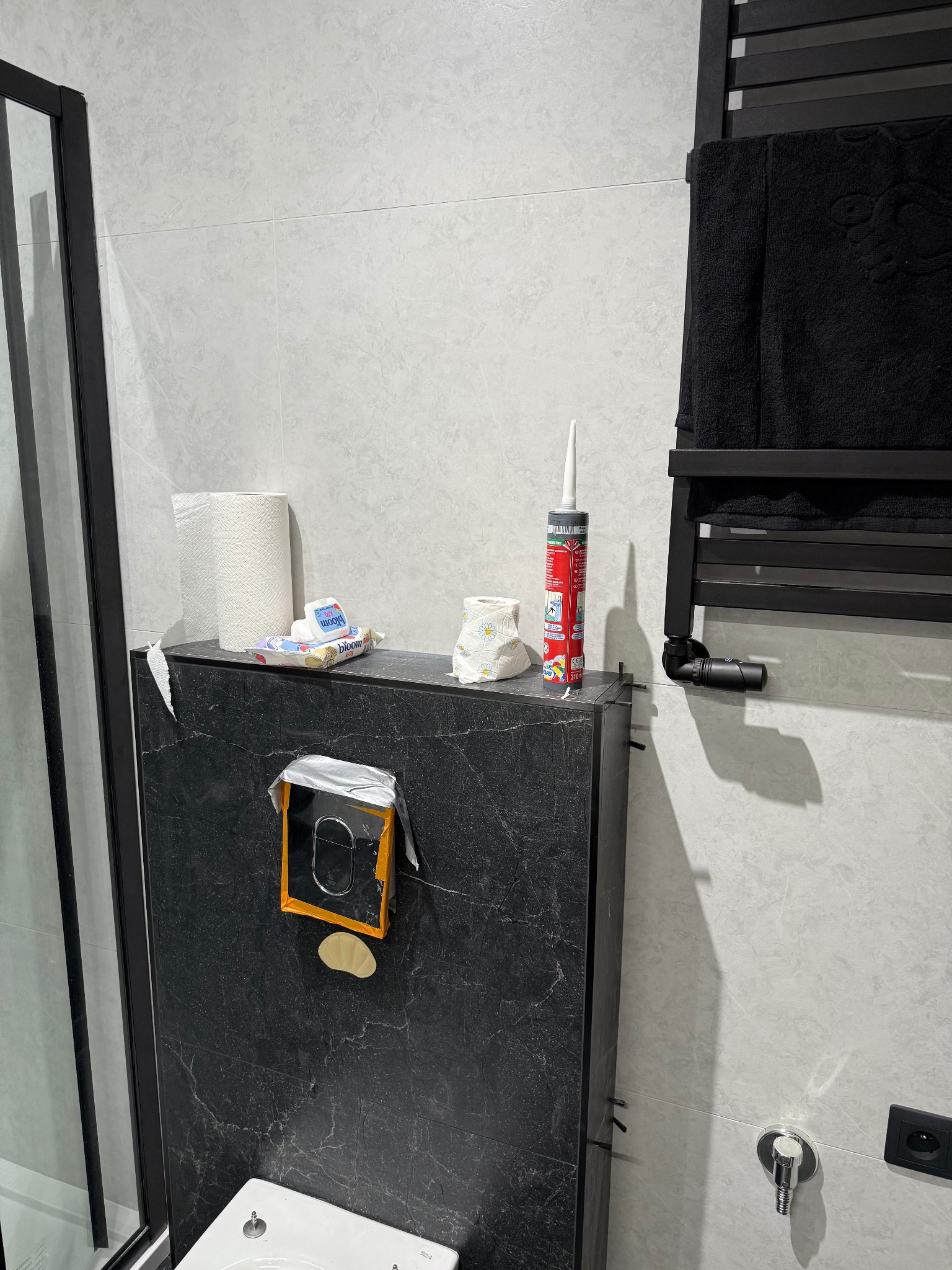 Wykończona łazienka we Wrocławiu: czarna zabudowa WC z marmurowym wzorem, szare płytki, czarny grzejnik drabinkowy z ręcznikiem i akcesoria łazienkowe.