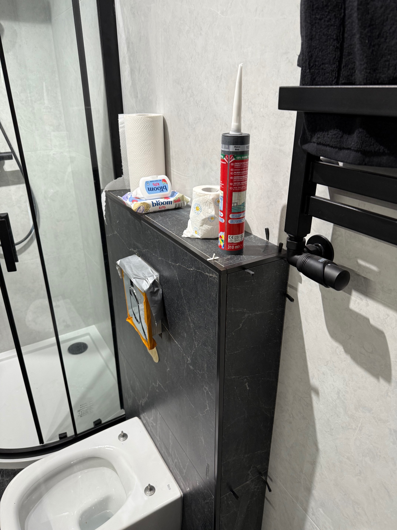 Wykończona łazienka z czarną armaturą i płytkami imitującymi marmur. Widoczna kabina prysznicowa, toaleta, grzejnik i akcesoria łazienkowe. Tubka silikonu na szafce.