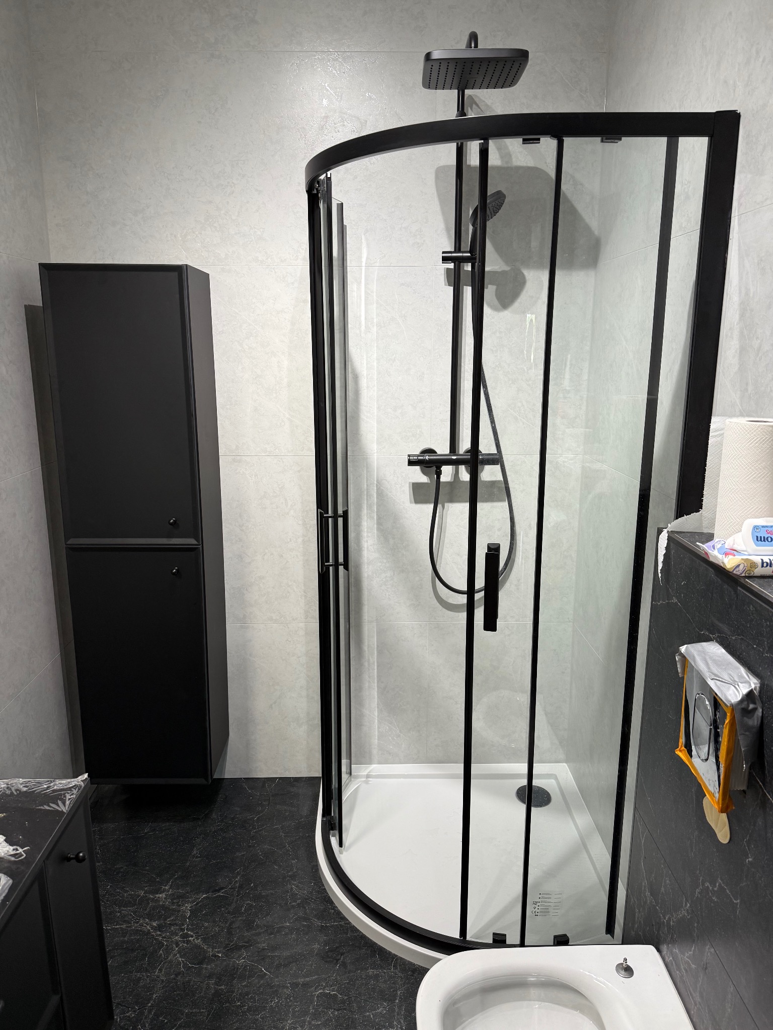 Nowoczesna łazienka z czarną kabiną prysznicową i szafką, jasne płytki na ścianach, ciemna podłoga z marmurkowym wzorem. Minimalistyczny design.