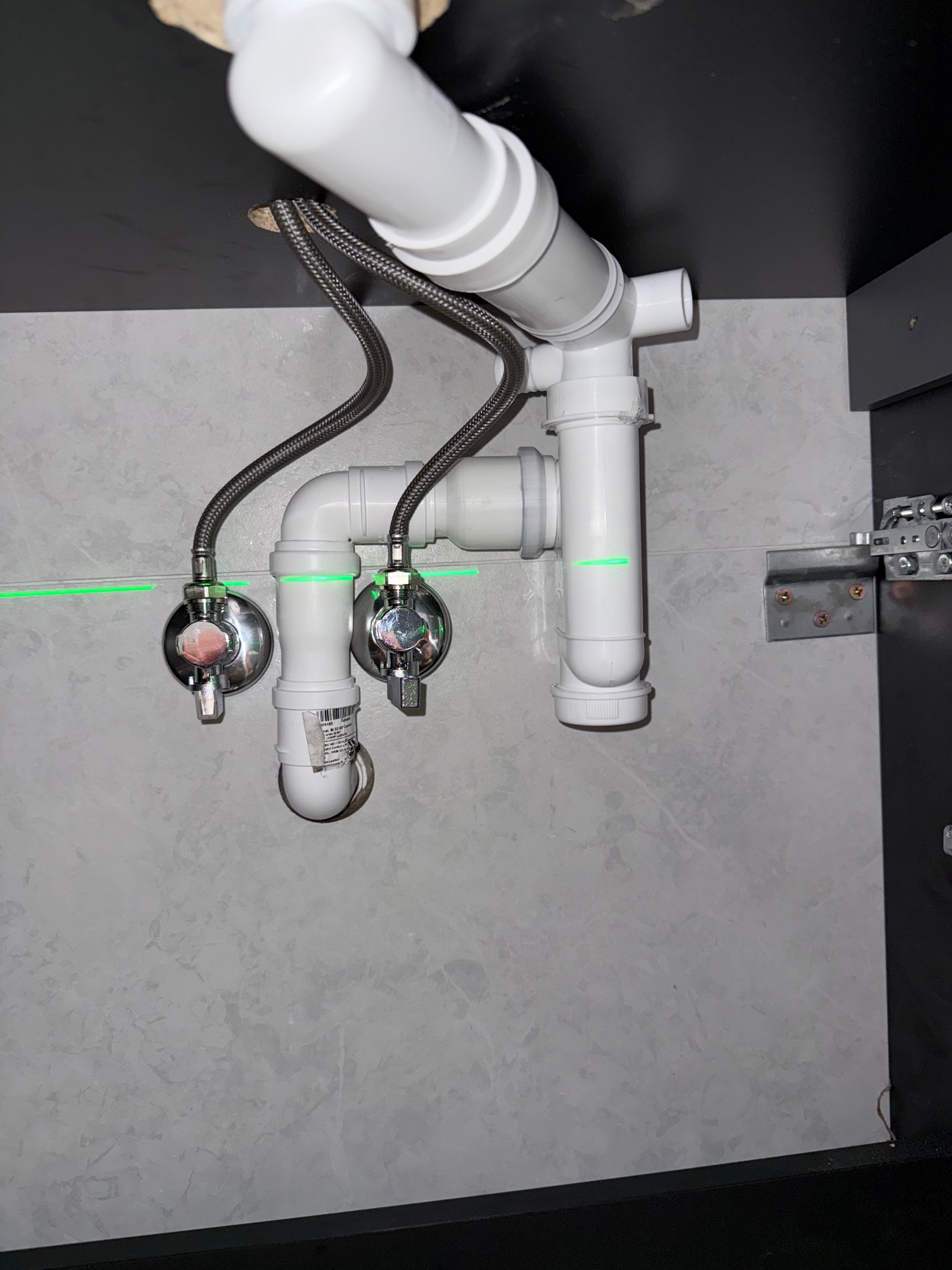 Zamontowana, biała instalacja odpływowa pod umywalką, widoczne zawory z wężykami w oplocie, poziomica laserowa, szare płytki.