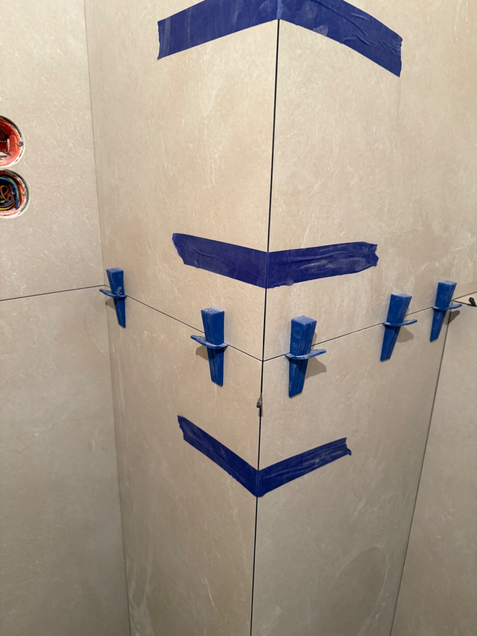 Układanie jasnych płytek w narożniku z użyciem niebieskiej taśmy i klipsów poziomujących we Wrocławiu. Widoczne gniazdka elektryczne w ścianie.