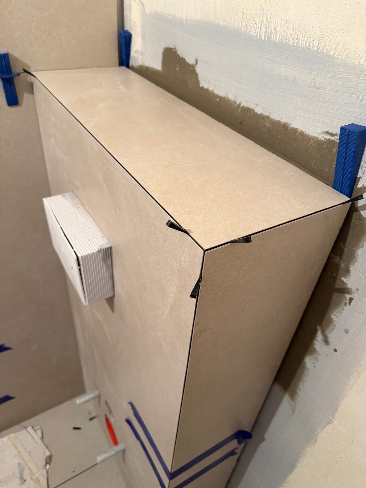 Fragment obudowy z płytek w trakcie montażu, z użyciem poziomujących klinów i krzyżyków dystansowych, widoczne surowe ściany w tle.