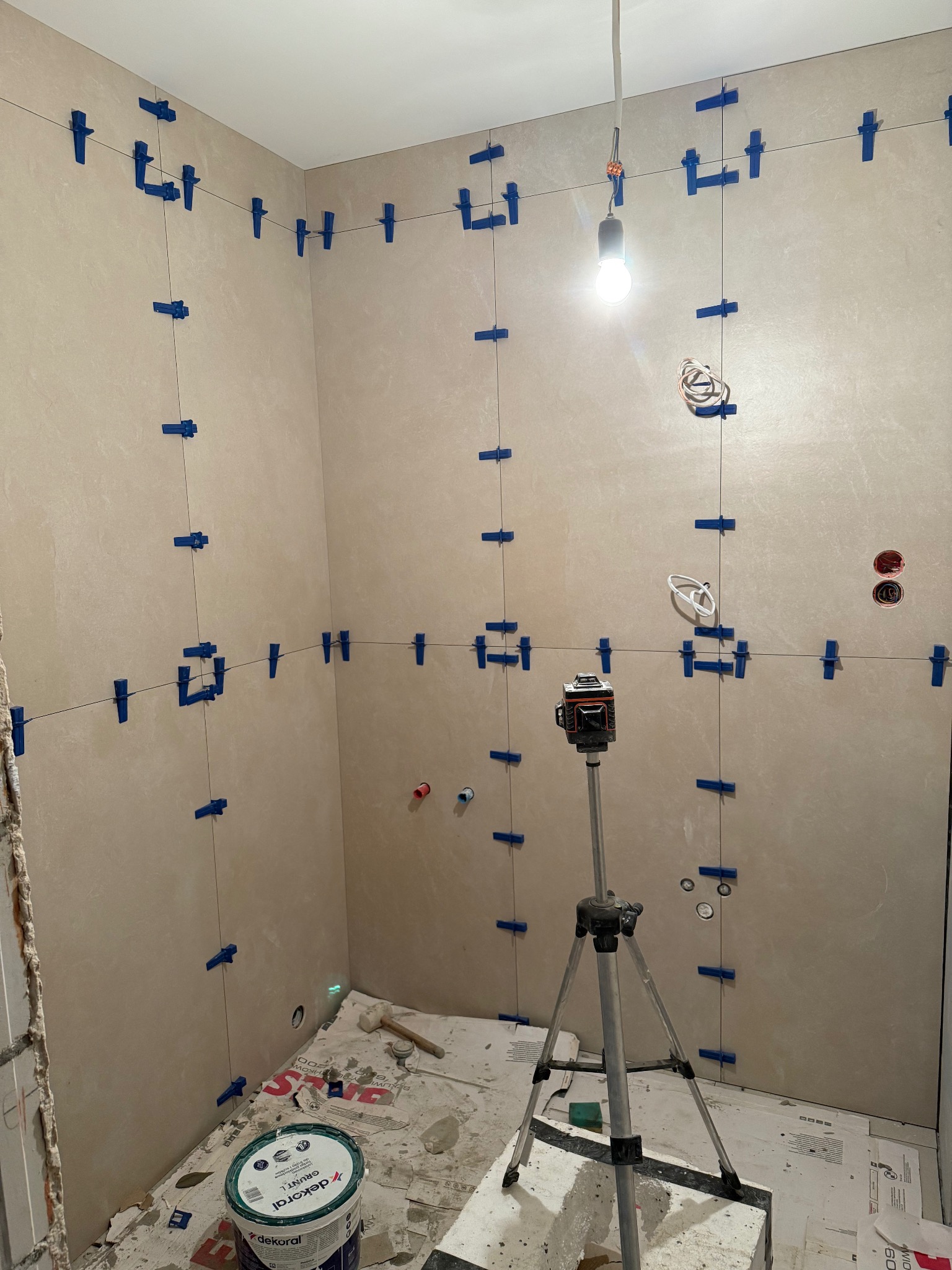 Układanie płytek w łazience z użyciem poziomicy laserowej i systemu poziomowania płytek. Widoczne narzędzia i materiały budowlane. Jasne płytki na ścianach.