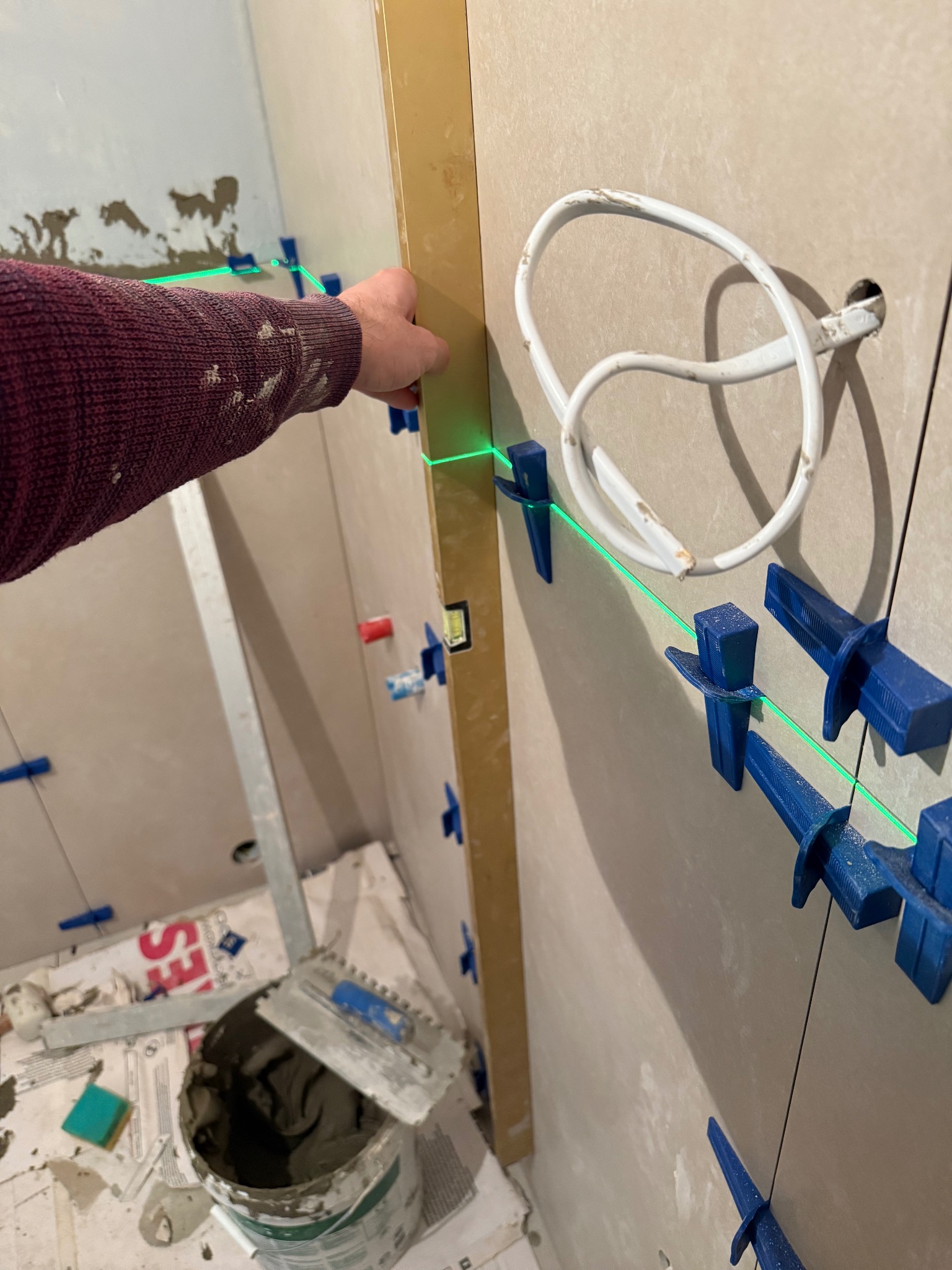 Układanie płytek na ścianie z użyciem poziomicy laserowej i klipsów. Widoczny fragment ręki w swetrze, wiadro z klejem i narzędzia glazurnicze.