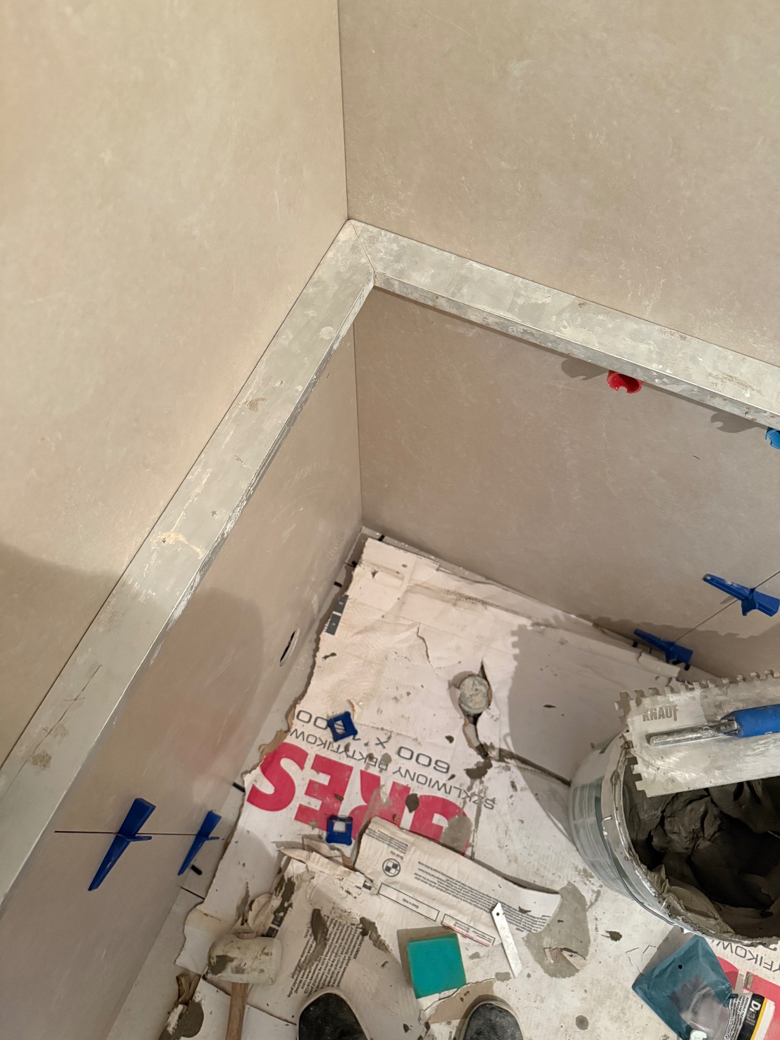 Wnętrze łazienki w trakcie prac glazurniczych: świeżo położone, beżowe płytki ścienne, poziomice, krzyżyki dystansowe i wiadro z zaprawą klejową.