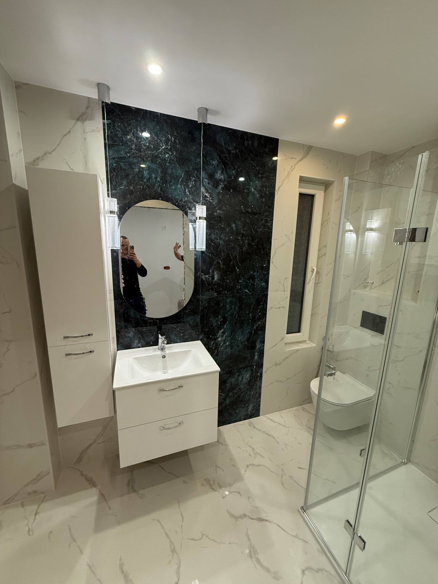 Wykończona łazienka z marmurowymi płytkami, kabiną prysznicową i umywalką z szafką. Na ścianie dekoracyjny panel w odcieniach zieleni i czerni. W lustrze odbicie osoby.