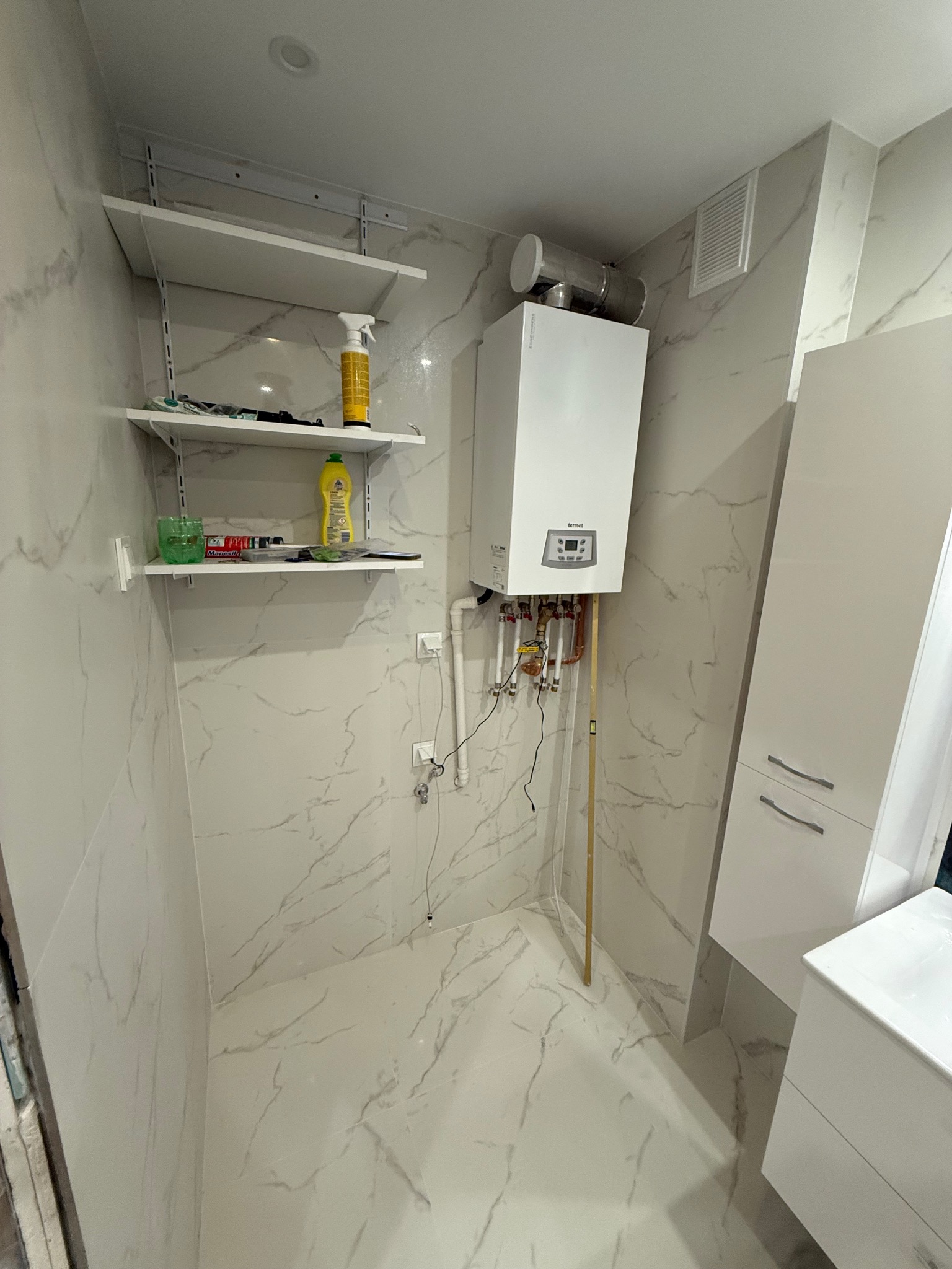 Biała łazienka z marmurowymi płytkami, wiszącym piecem gazowym Termet, półkami i szafką pod umywalką. Widoczne rury i instalacja. Nowoczesny design.