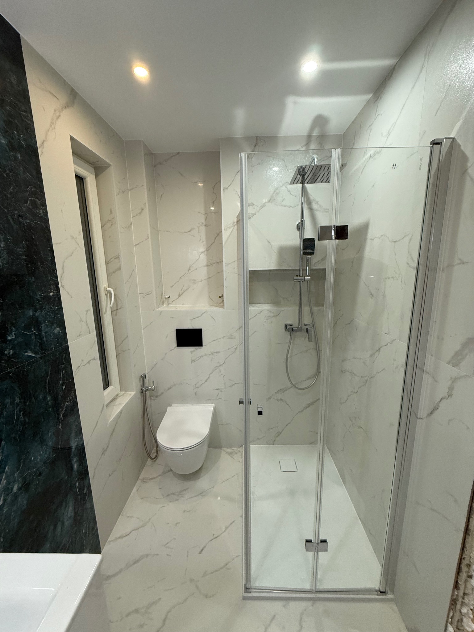 Nowoczesna łazienka z kabiną prysznicową typu walk-in, białe płytki imitujące marmur, podwieszana toaleta i akcent z ciemnego kamienia na ścianie.