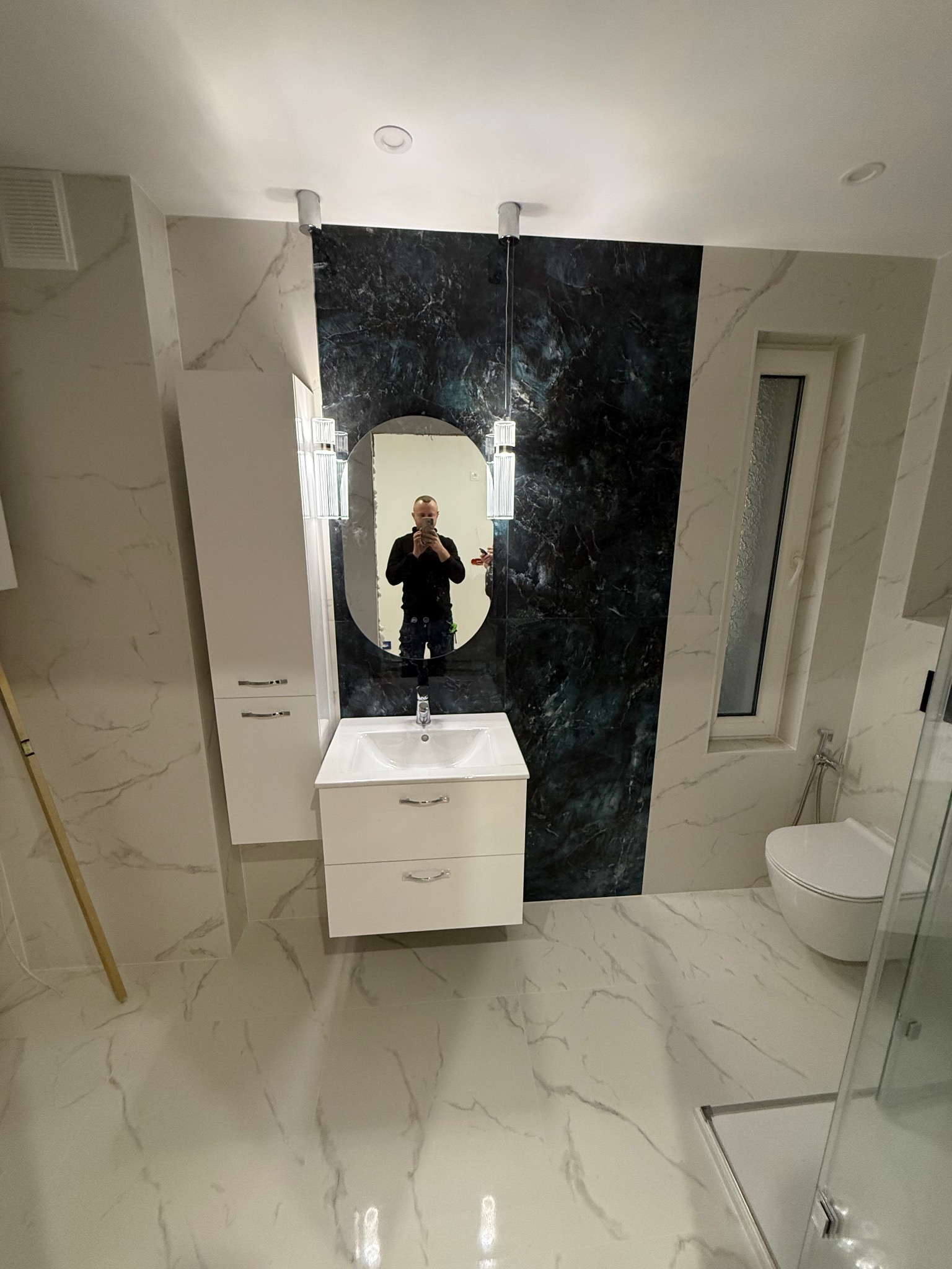 Nowoczesna łazienka z marmurowymi płytkami, owalnym lustrem i ciemnozieloną ścianą. Widoczna toaleta i kabina prysznicowa. W lustrze odbija się osoba robiąca zdjęcie.