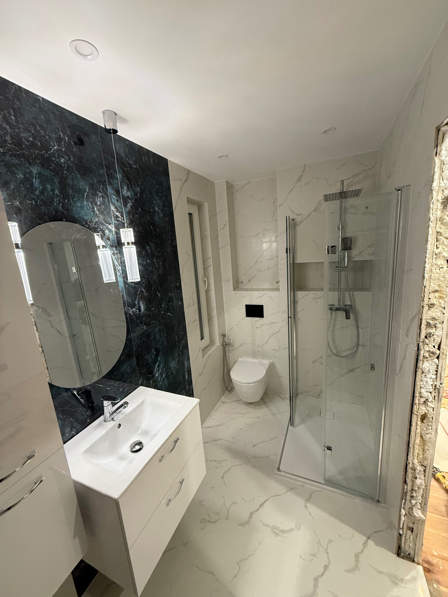 Nowoczesna łazienka z kabiną prysznicową, umywalką z szafką i toaletą. Marmurowe płytki na podłodze i ścianach, akcent z ciemnego marmuru za lustrem.