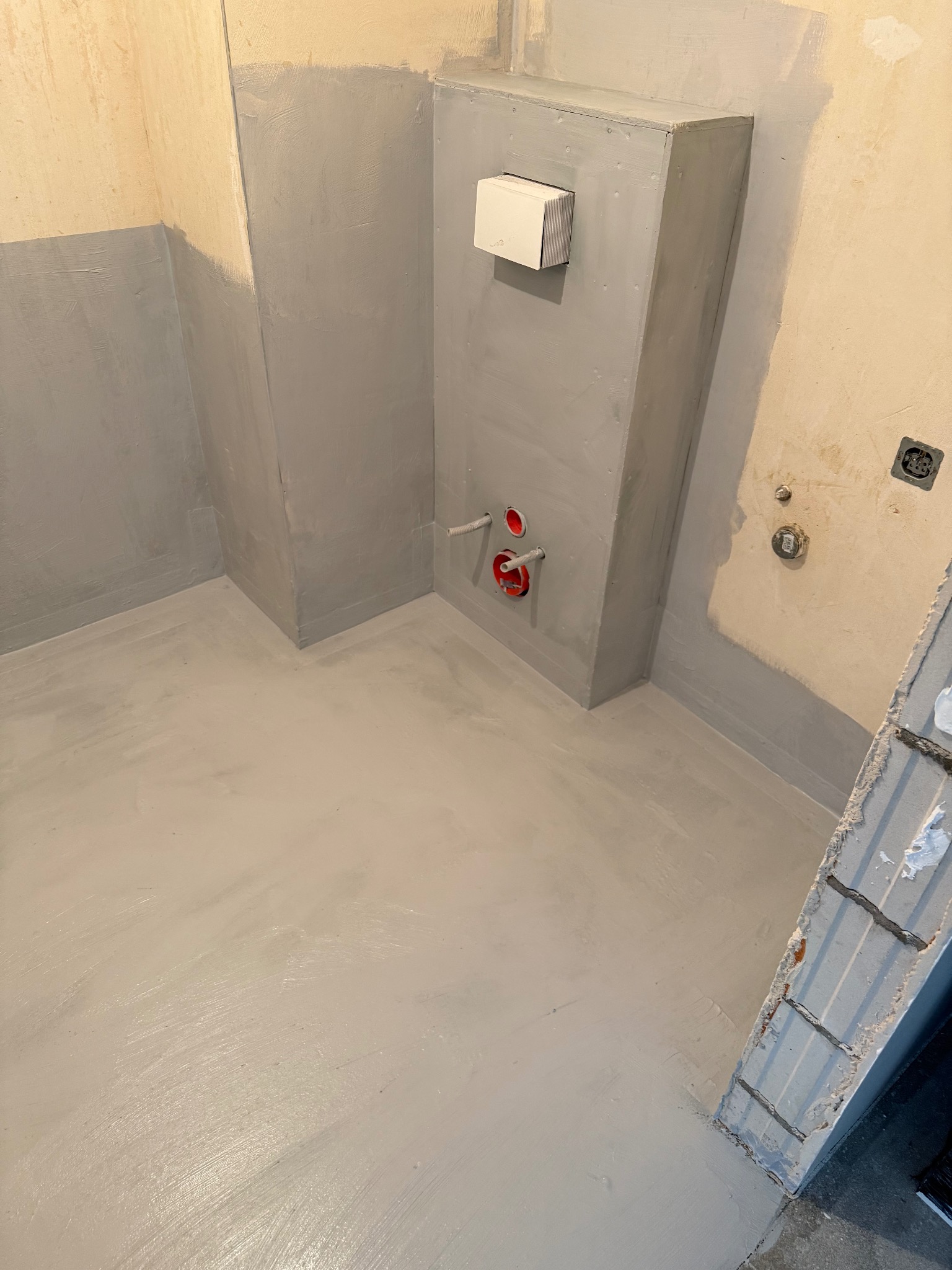 Szara hydroizolacja w łazience w trakcie remontu. Widoczne zabezpieczone ściany, podłoga i zabudowa stelaża WC. Przygotowanie do dalszych prac wykończeniowych.