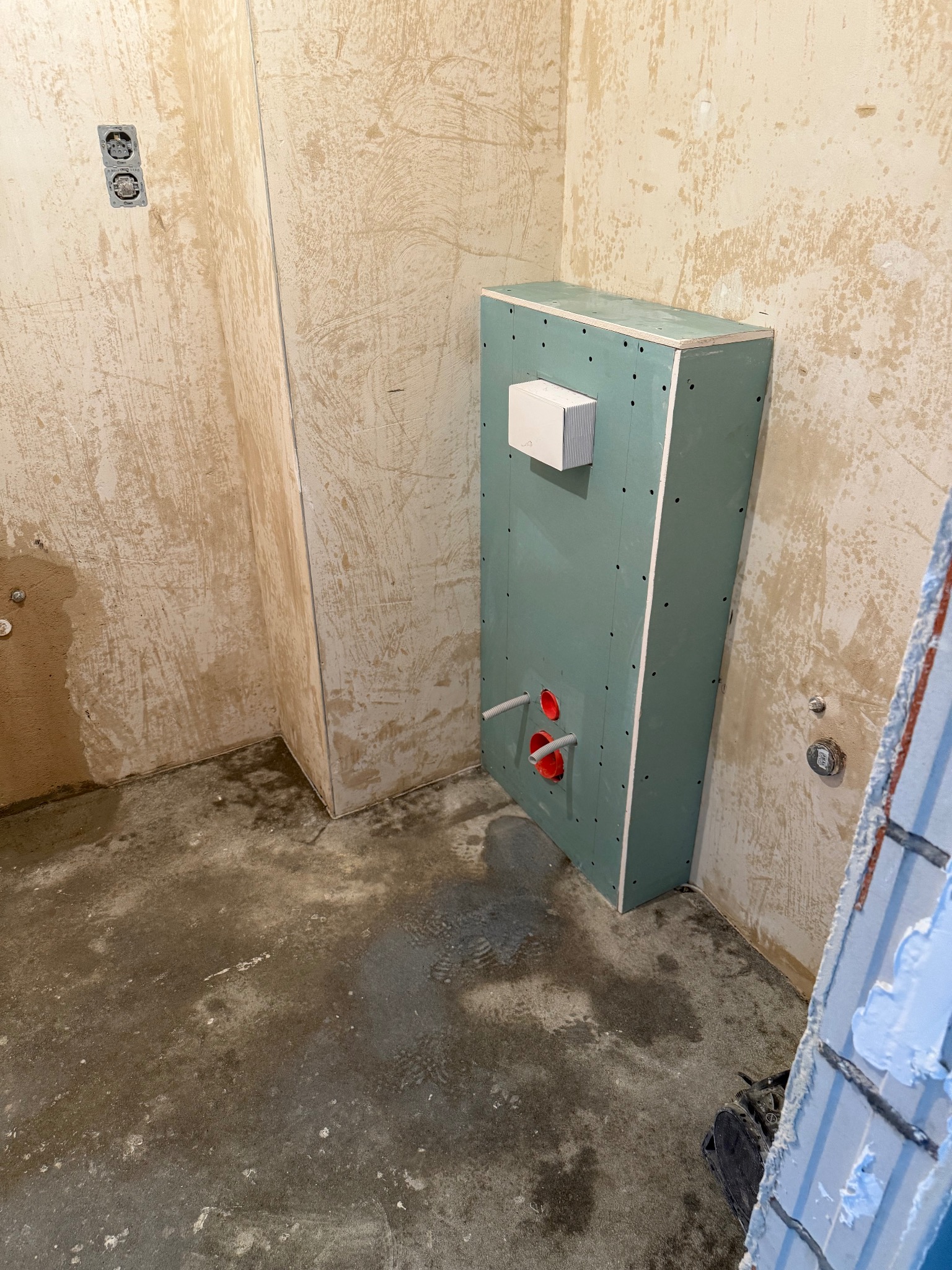 Narożnik łazienki w trakcie prac: surowe ściany z gładzią, zabudowa z płyty G-K z instalacją hydrauliczną, widoczne gniazdka elektryczne i fragment układanych płytek.