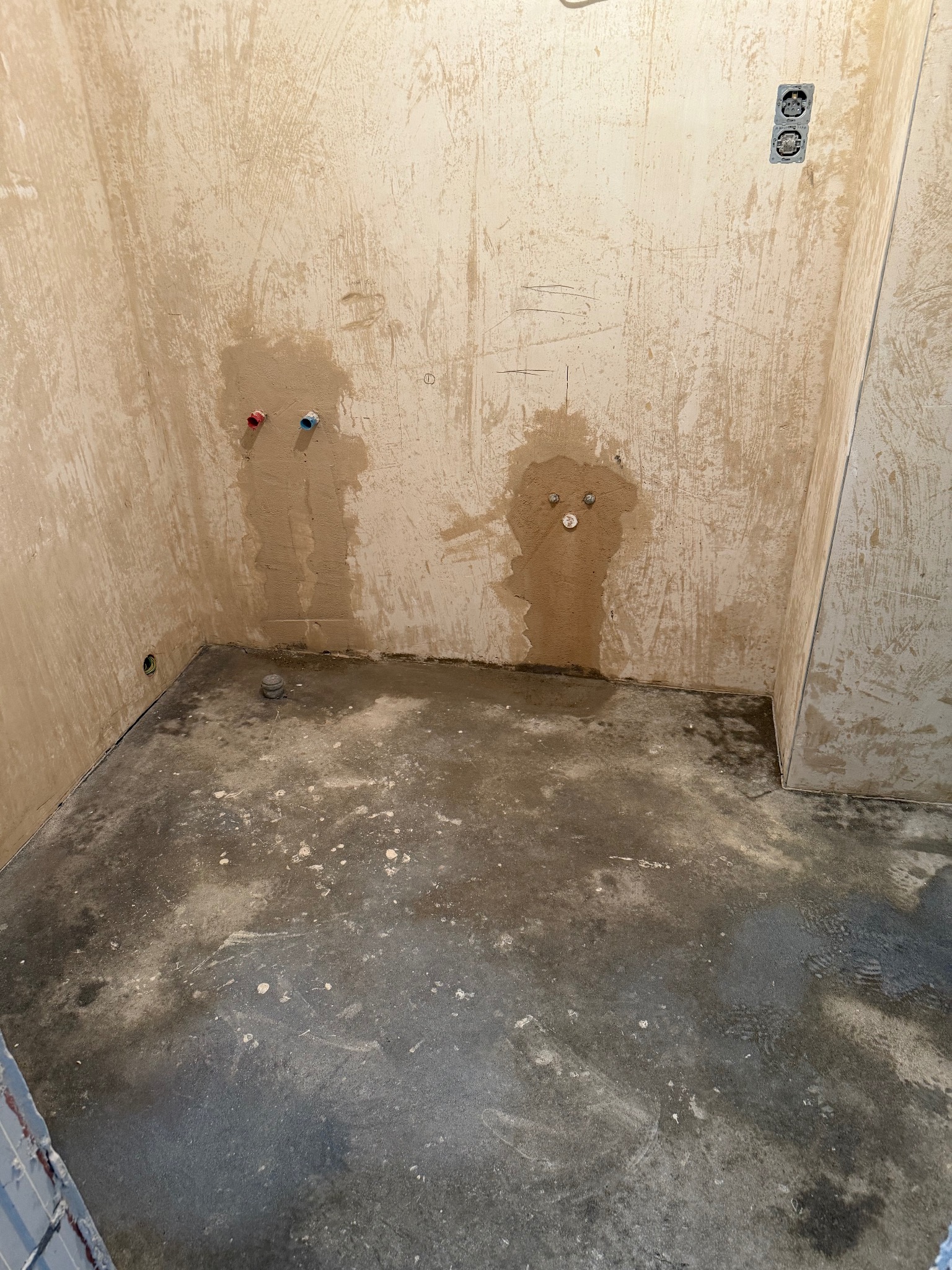 Surowe wnętrze łazienki w trakcie remontu: ściany z resztkami starej farby, widoczne przyłącza wodne i elektryczne, surowy beton na podłodze z plamami farby.