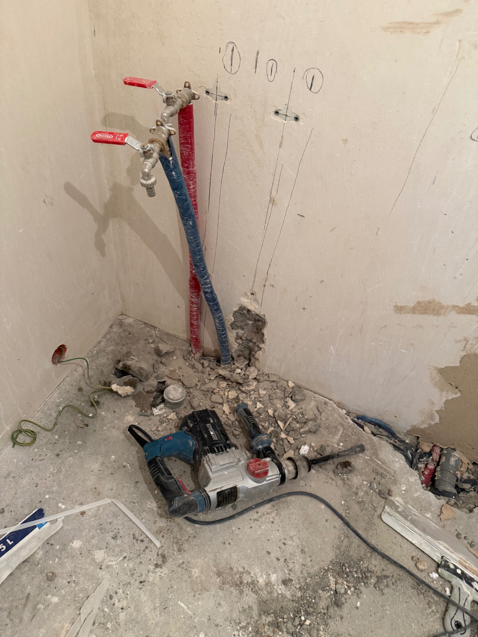Remont łazienki w trakcie: instalacja wodna z zaworami, kable elektryczne, ślady po kuciu w ścianie, gruz i wiertarka udarowa na podłodze. Widok na narożnik pomieszczenia.