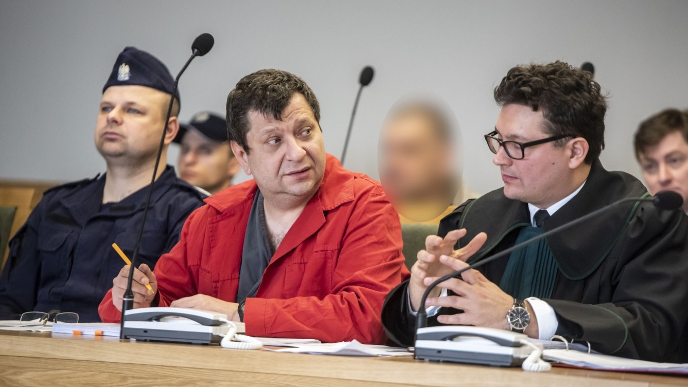 Sala sądowa: mężczyzna w czerwonej kurtce i adwokat w todze podczas rozprawy, obok policjant w mundurze. Mikrofony i telefony na stole. Rozprawa sądowa w toku.