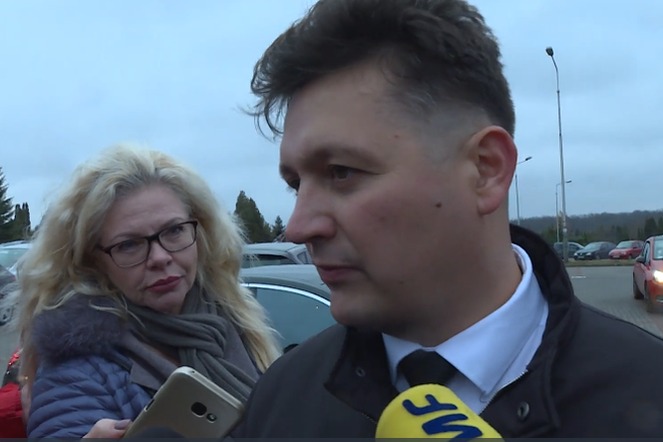 Wywiad z prawnikiem: Mężczyzna w garniturze odpowiada na pytania dziennikarzy z mikrofonami, w tle kobieta z telefonem.